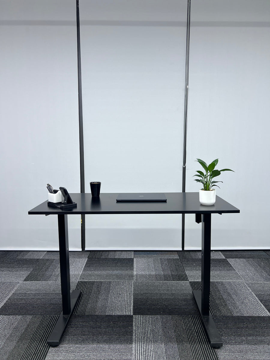 Black adjustable desk in an office setting with a plant and cup on it.