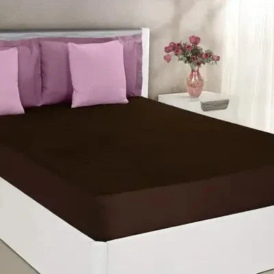 Brown fitted sheet on a bed with purple pillows and a vase of flowers on a nightstand.