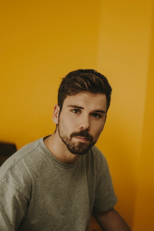 Man with a beard and short hair wearing a gray t-shirt against a yellow wall.